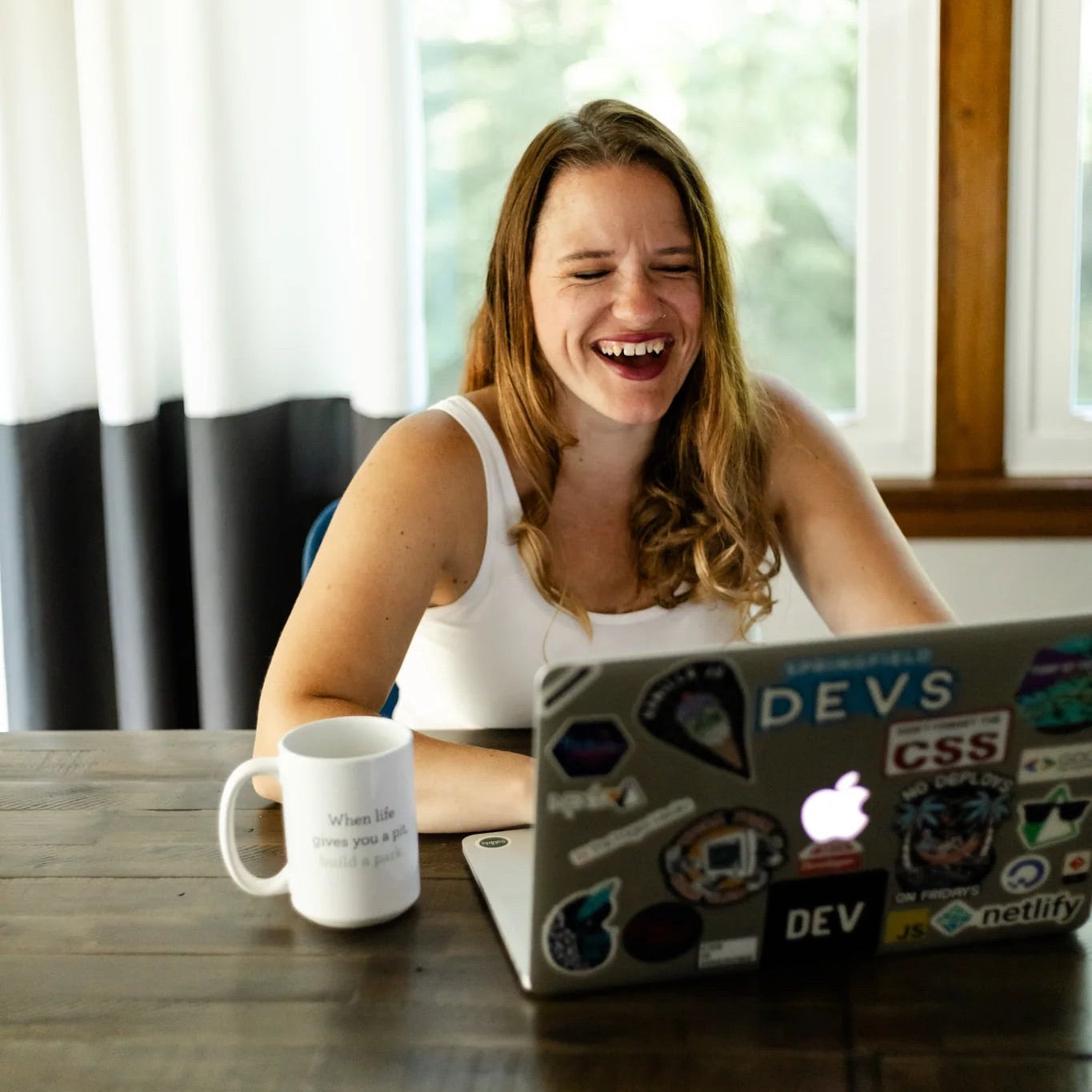 Kendra laughing at a laptop with stickers on a wooden table