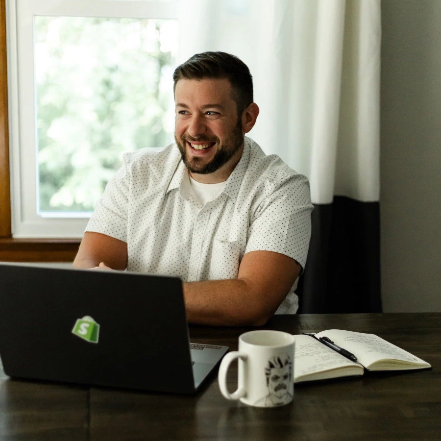 Taylor sitting at a desk with a laptop, mug, and notebook in a bright room with a window.